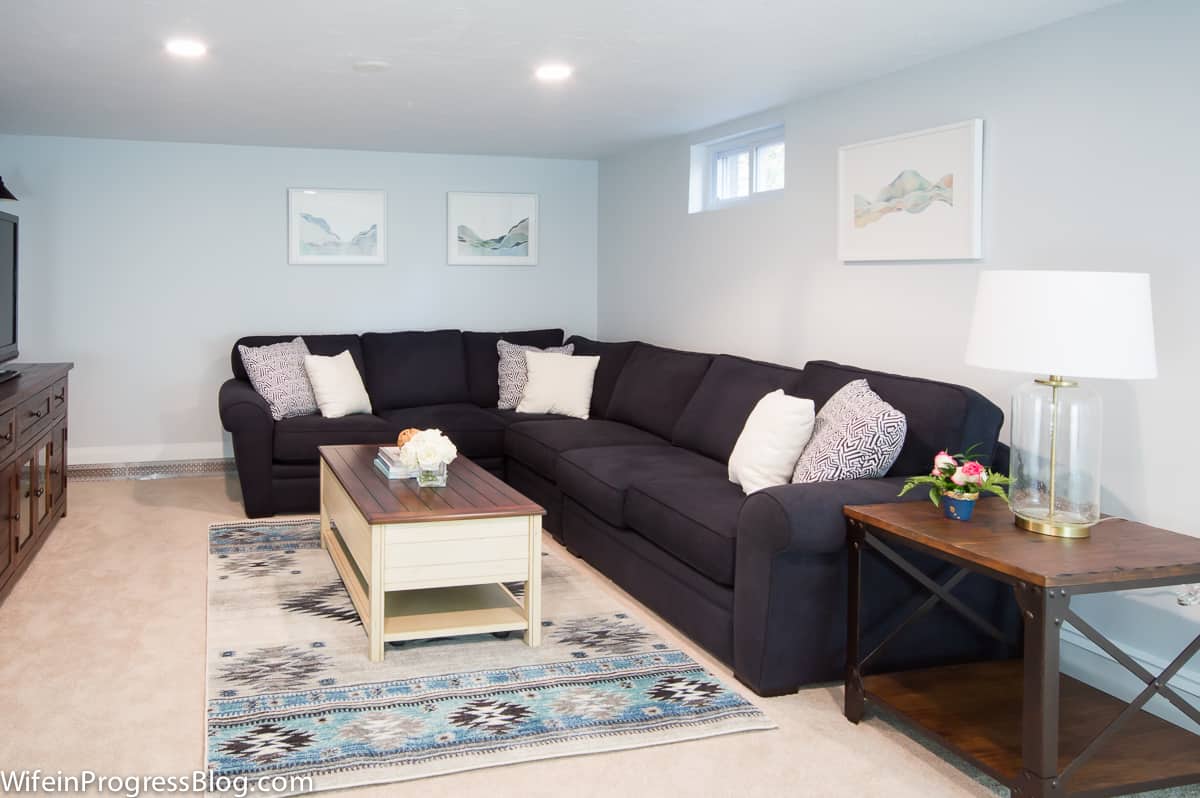 A living room with a dark sectional sofa with throw pillows, a rectangular area rug on beige carpeting and a beige coffee table