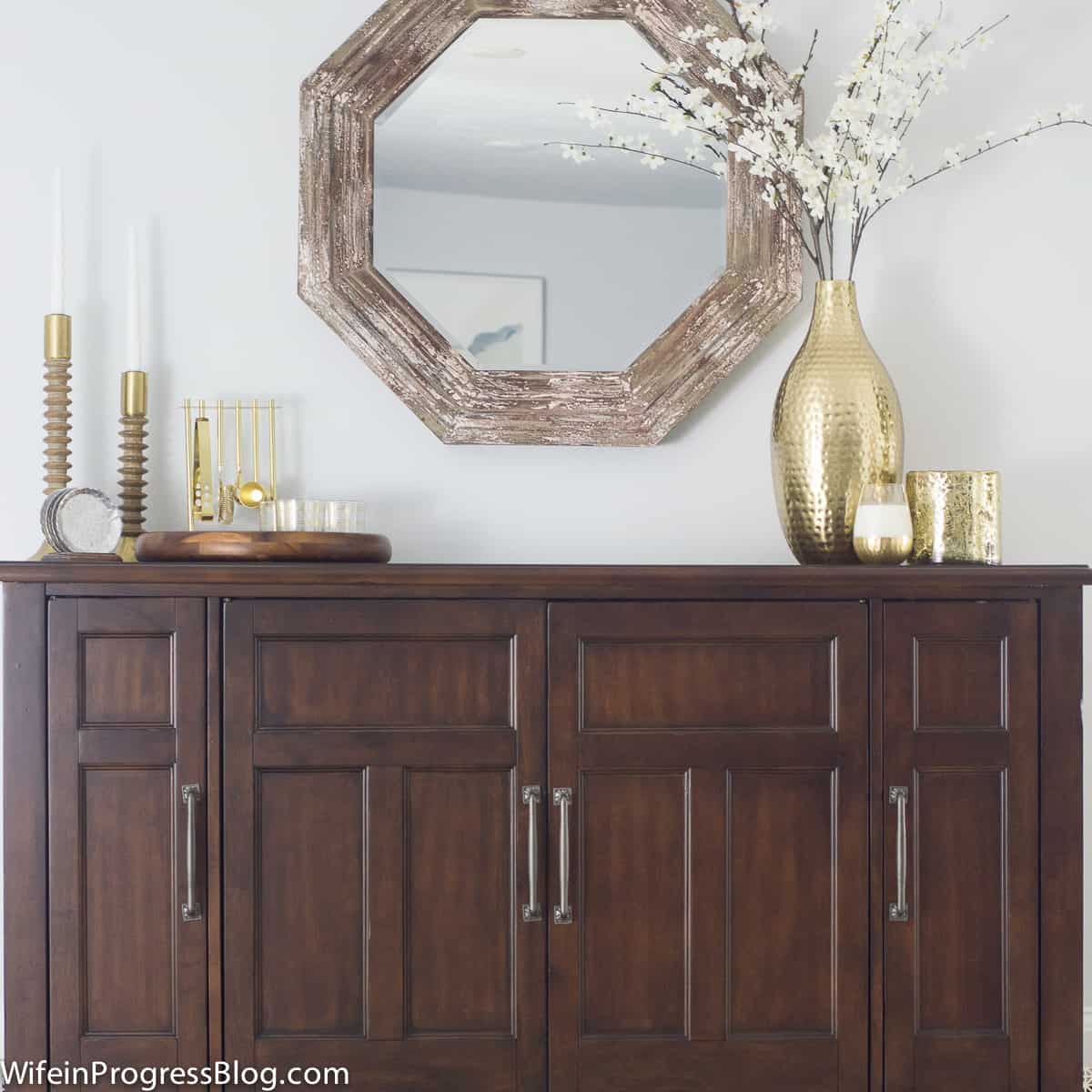 A large side table with various gold decor items on top and a hexagonal mirror above in a rustic, wood frame