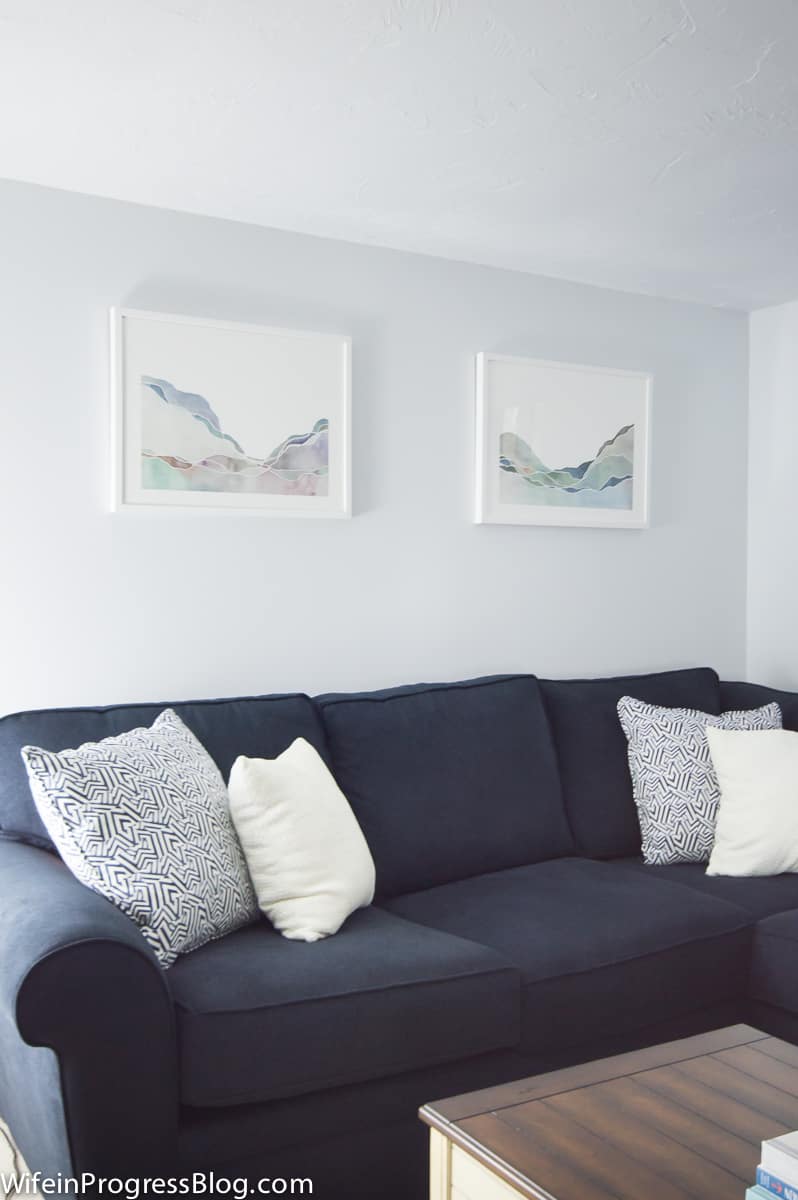 A navy sofa with white and blue throw pillows and two landscape drawings in white frames on the wall