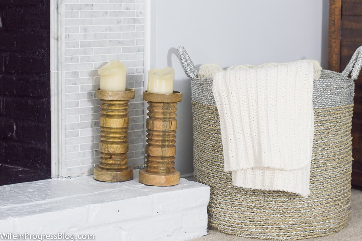 The corner of a fireplace hearth, holding two sturdy wooden candlesticks with beige candles, next to a wicker basket with a blanket draping over the side