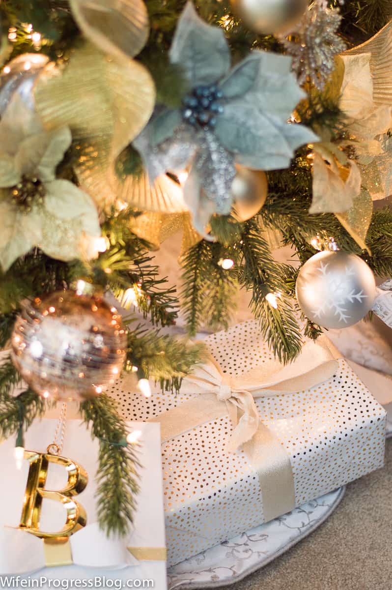 The base of a lit Christmas tree, with gold and light blue poinsettia flowers, copper and silver ornament balls and a large, wrapped gift below