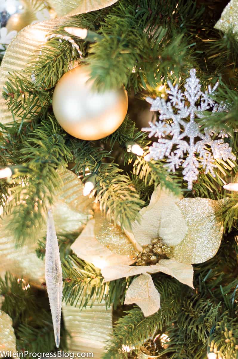 A close up of a golden poinsettia, white snowflake and golden ornament balls on a lit Christmas tree