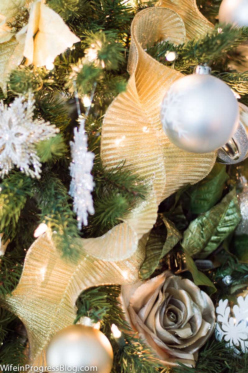 A close up of gold ribbon trailing down a Christmas tree, with silver ornament balls and white snowflakes nearby