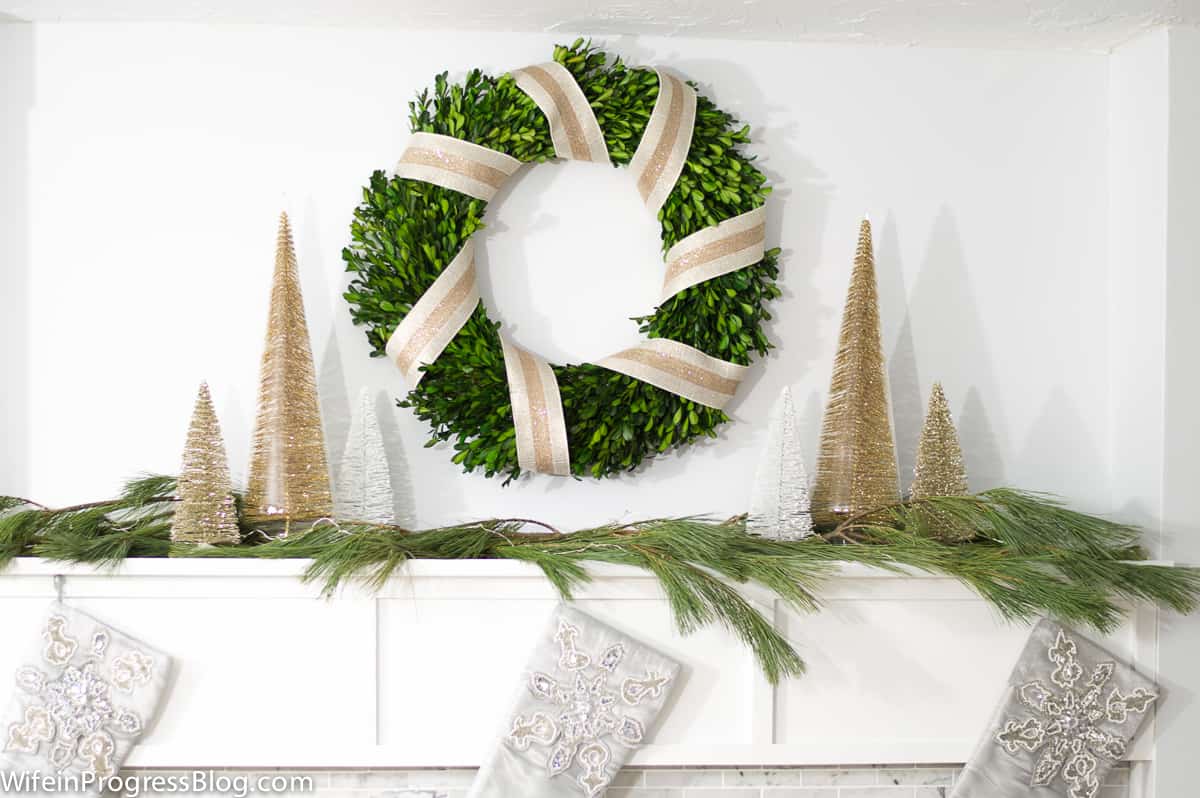 A green wreath, wrapped in gold ribbon, above a mantel holding pine branches and gold cones resembling trees