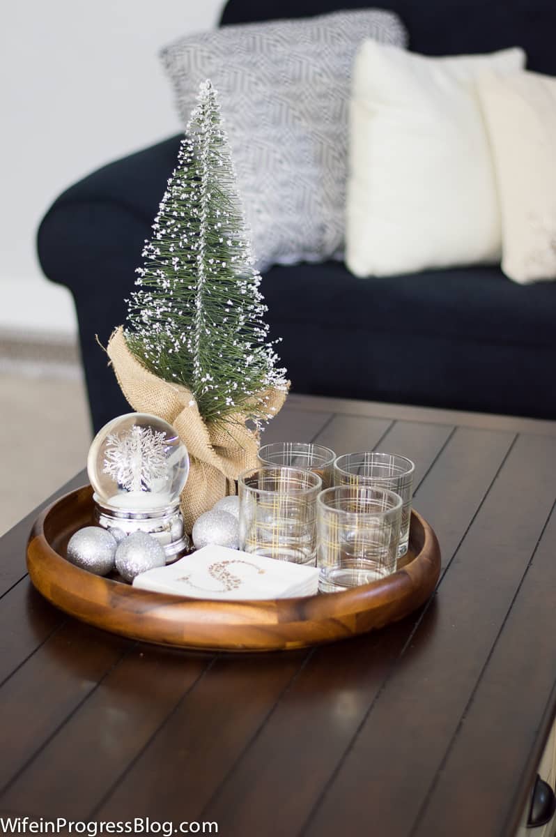 A round, wooden tray on a coffee table holding a pine tree in burlap, a silver snow globe, silver ornaments and four glass tumblers