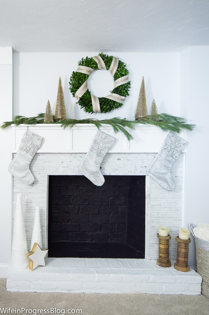 Three grey stockings hung on a fireplace mantel, with a green wreath above, wrapped in gold ribbon
