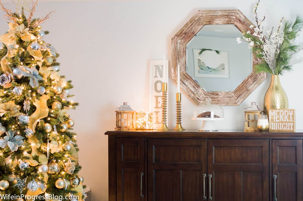 A dark, wooden side table with a hexagonal mirror and various holiday decor items on top, next to a lit Christmas tree
