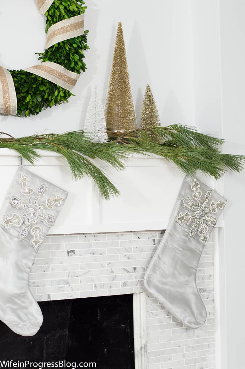 Grey stockings with bead embellishments on top, hung from mantel decorated with pine branches, gold trees and a green wreath