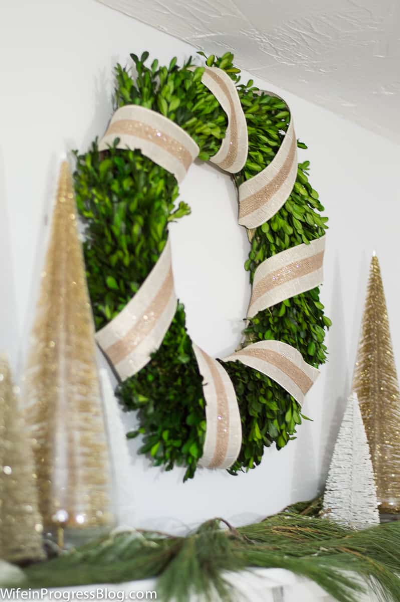 A close up of a green wreath with beige and gold striped ribbon wrapped around it, with tall, gold trees nearby
