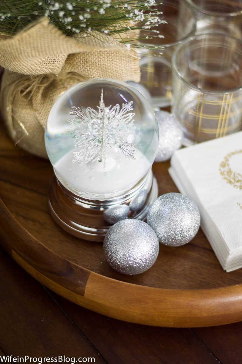 A close-up of a wooden tray with a silver snow globe, silver ornaments and napkins with the initial \'S\' on them, near glass tumblers