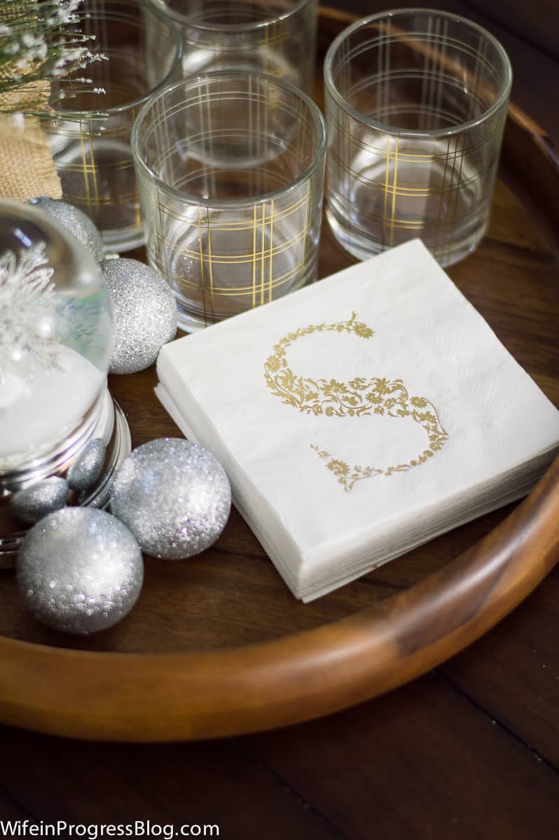 A close-up of cocktail napkins with the initial \'S\' in gold filigree pattern, near glass tumblers with gold detailing