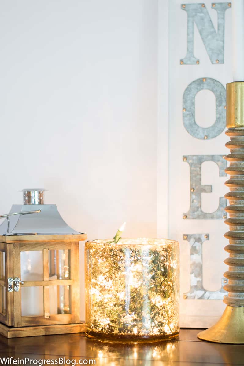 A lantern and a golden candle jar next to a tall, gold candlestick and a sign with the words \"Noel\"
