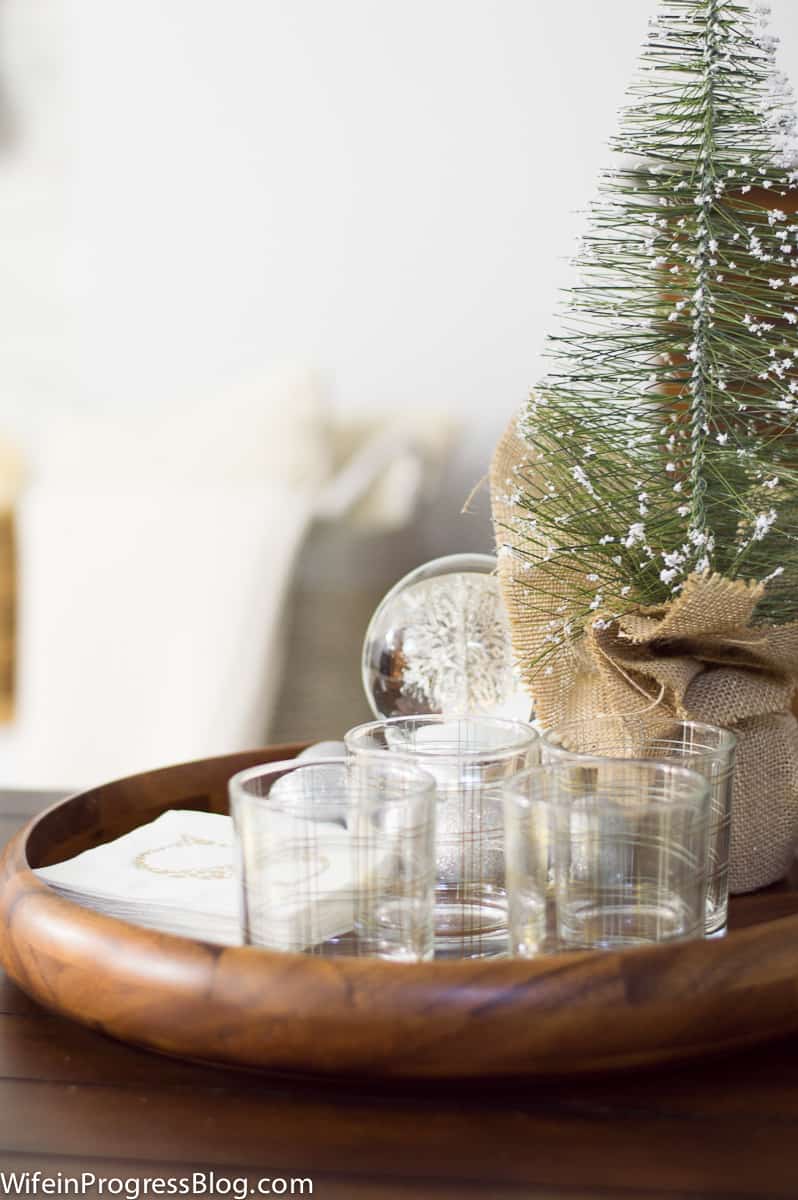 An artificial pine tree, wrapped in burlap, near glass tumblers in a round, wooden tray