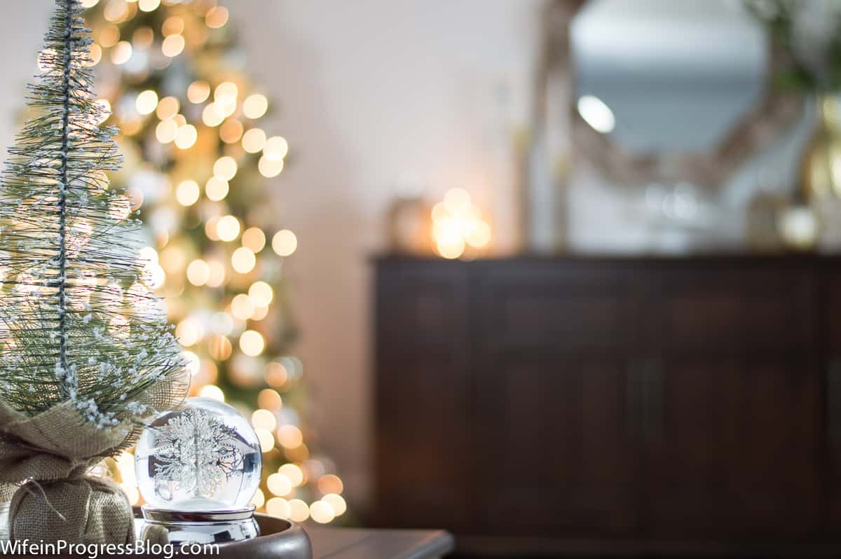 A small pine tree and a silver snow globe on a tray, with a glowing Christmas tree and a dark brown side table in the background