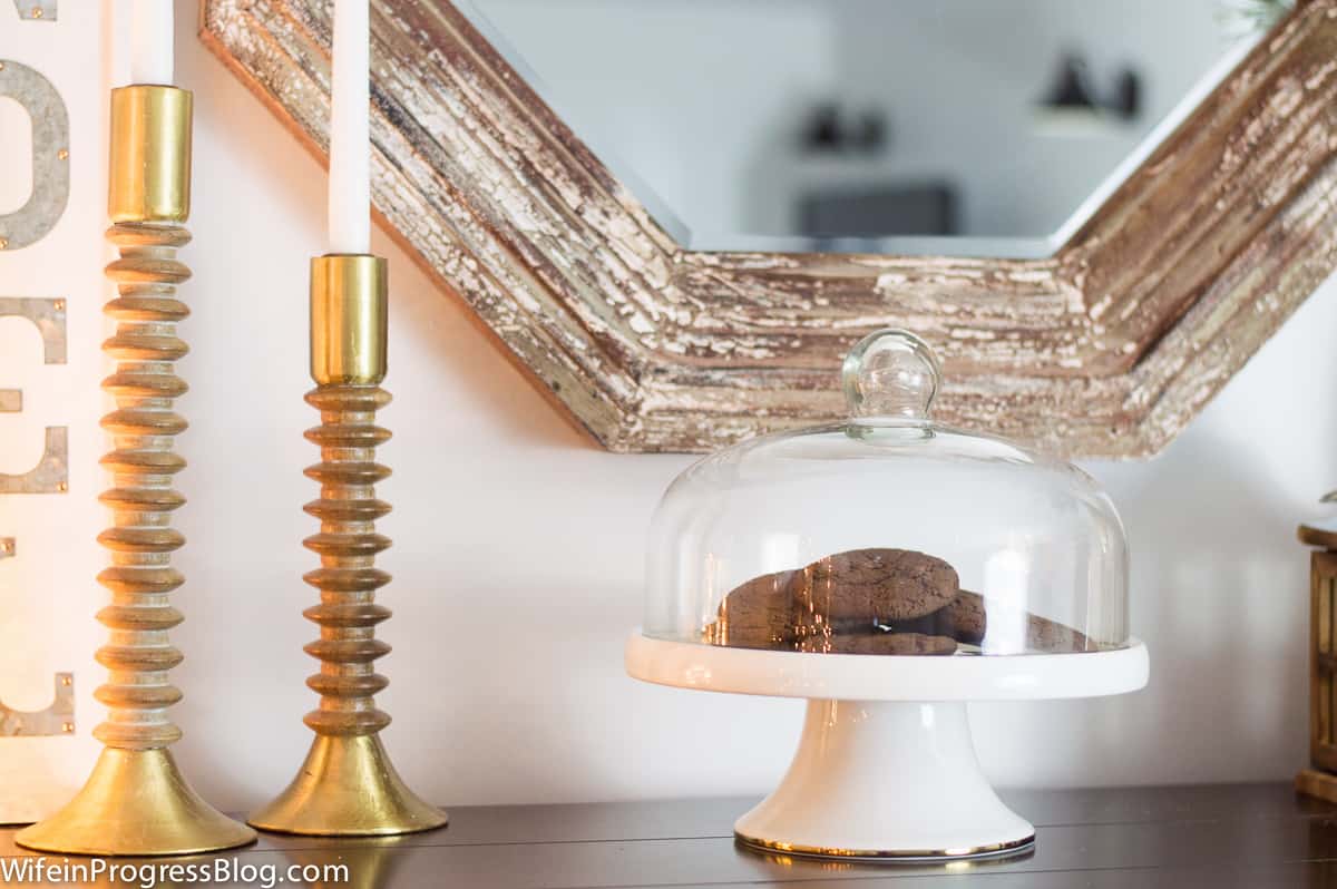 2 gold candlesticks to the left, a cake stand with glass dome cover and chocolate cookies underneath, with a hexagonal mirror nearby