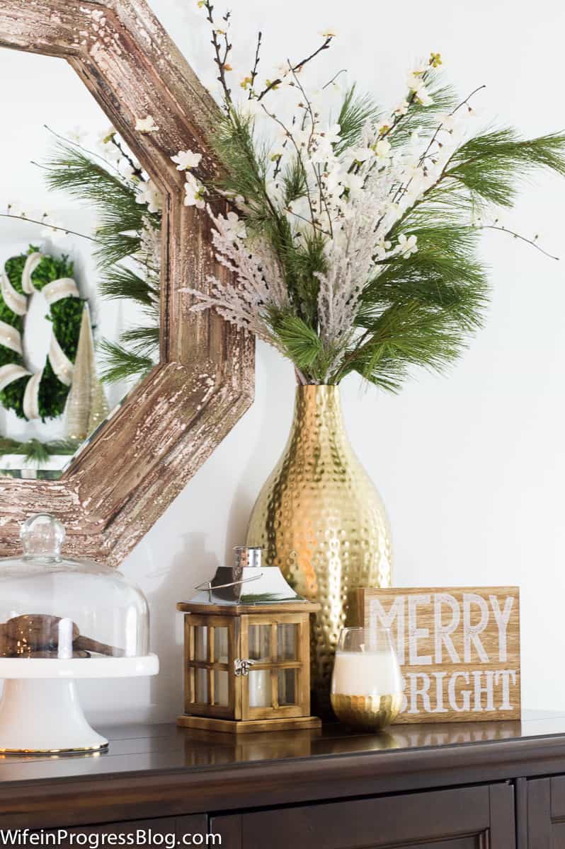 A tall, gold textured vase with a wide base and narrow opening, with winter greenery, near a mirror on the wall and various holiday decor beneath