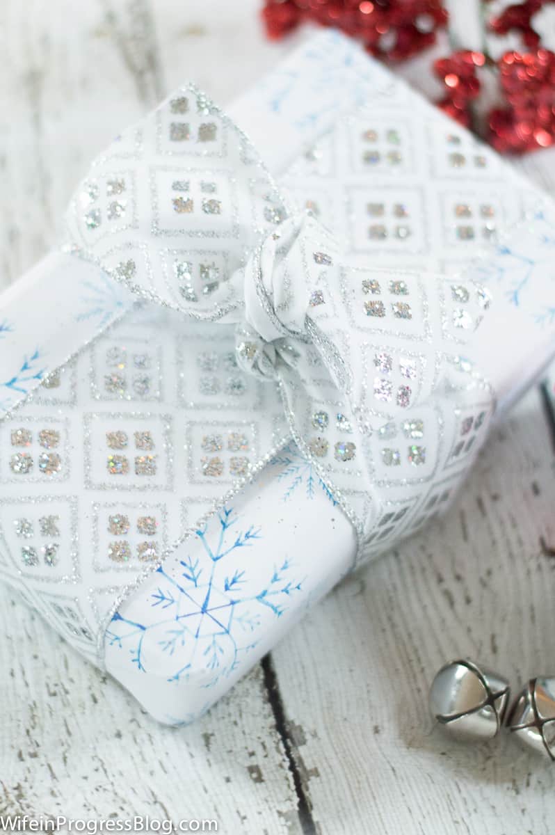 A rectangular gift wrapped in white wrapping with blue snowflake patterns, and tied with a wide, silver and white ribbon
