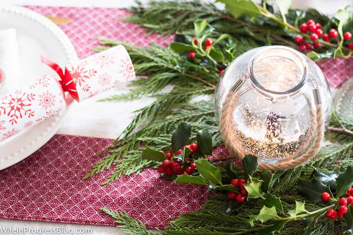 A short, round glass candle holder with gold detailing and a rope handle, resting on holly and green leaves on a red table runner