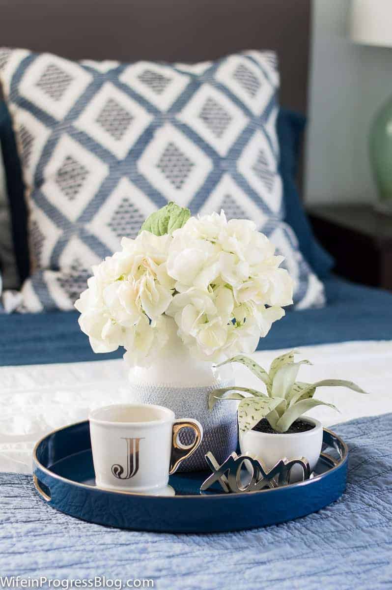 A round, blue tray on a bed, holding a gold/white mug with the initial \"J\", a blue vase with white flowers, and a small potted plant