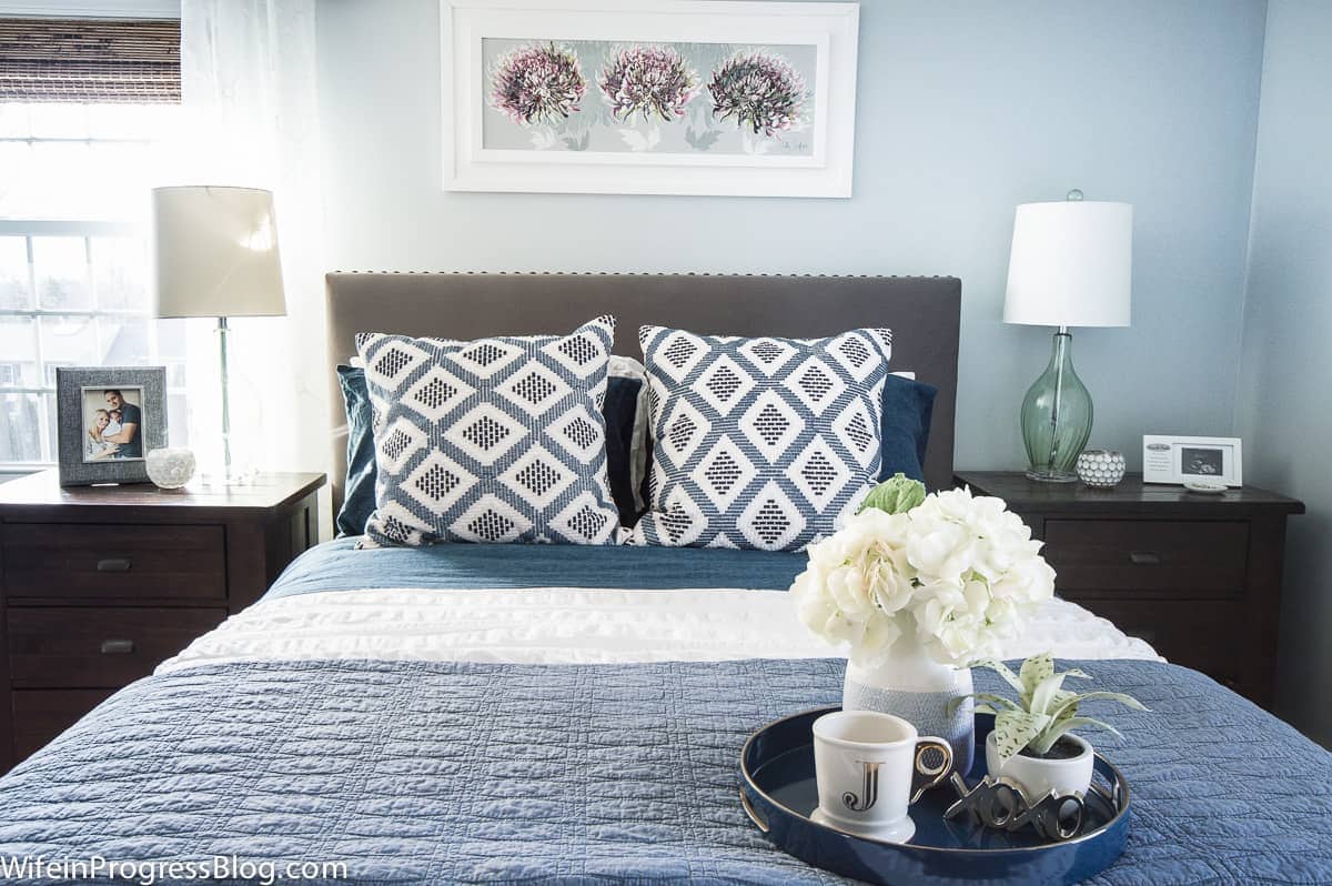 Rectangular white floral picture frame above bed with blue pillows & coverlet; round tray with floral arrangement and coffee mug nearby.