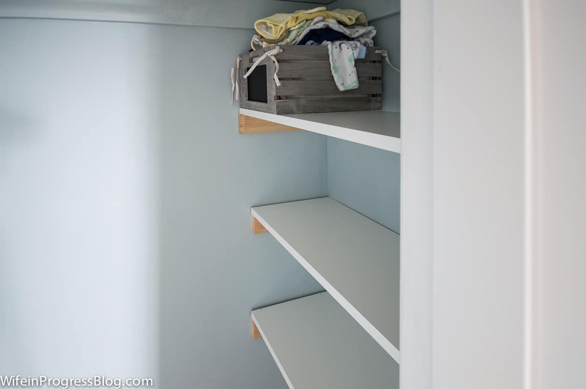 A trio of melamine shelves in the side of the closet, with unpainted cleats/braces supporting each shelf. A basket of baby bibs rests on top of one.