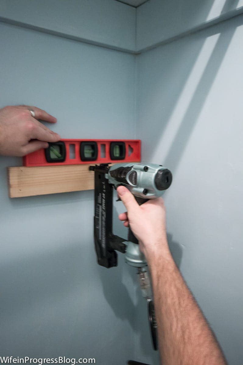 A person using a nail gun to secure a strip of wood to the wall, with a level above