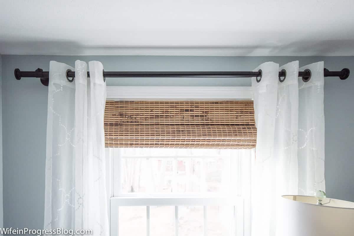 A top view of a window with a black curtain rod, white sheer grommet curtains and rolled up bamboo shades