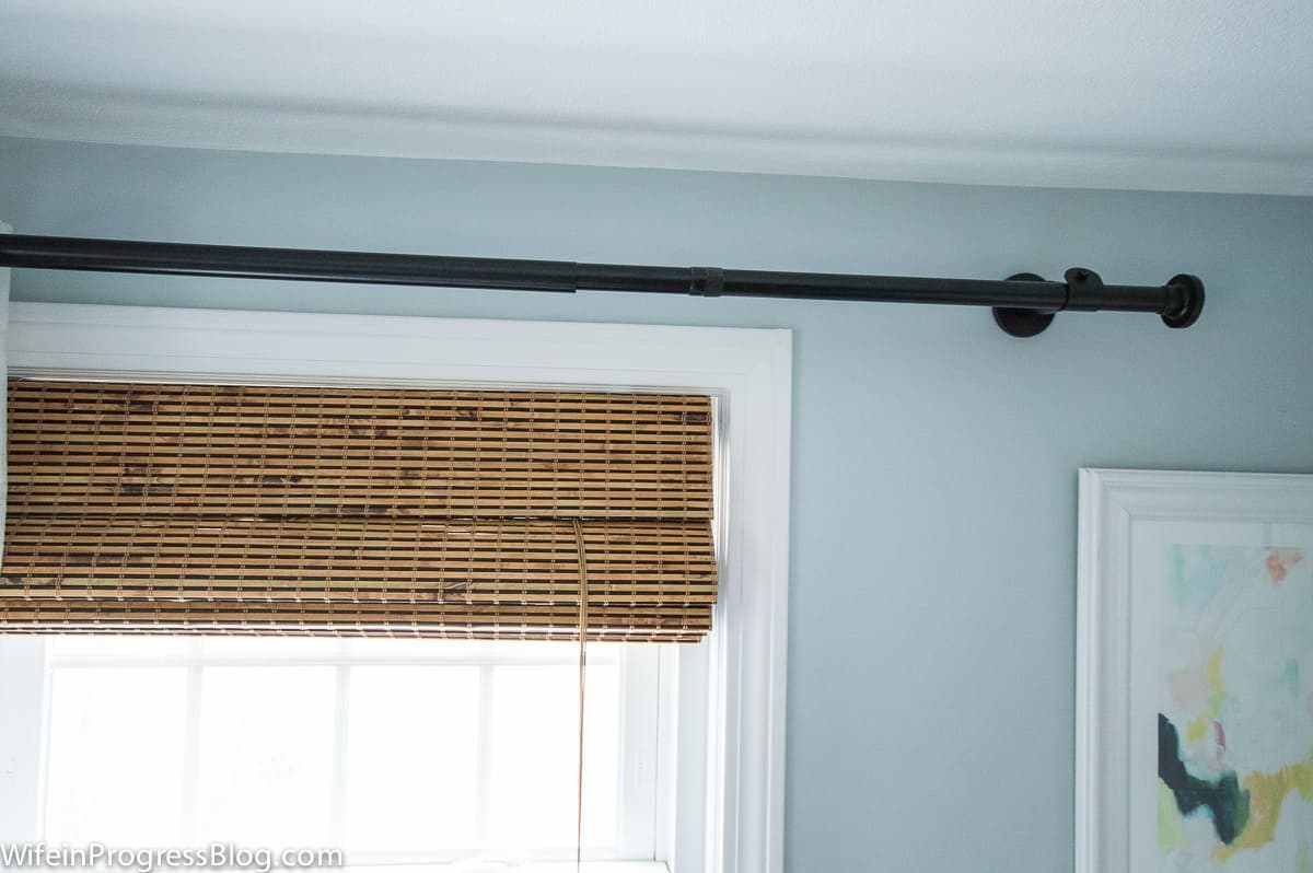 A close up of the top of a window, showing a black curtain rod and rolled-up bamboo blinds