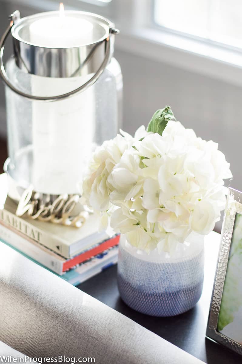 A narrow table behind the sofa, with a vase of flowers, a photo frame, and a glass candle holder on a stack of books