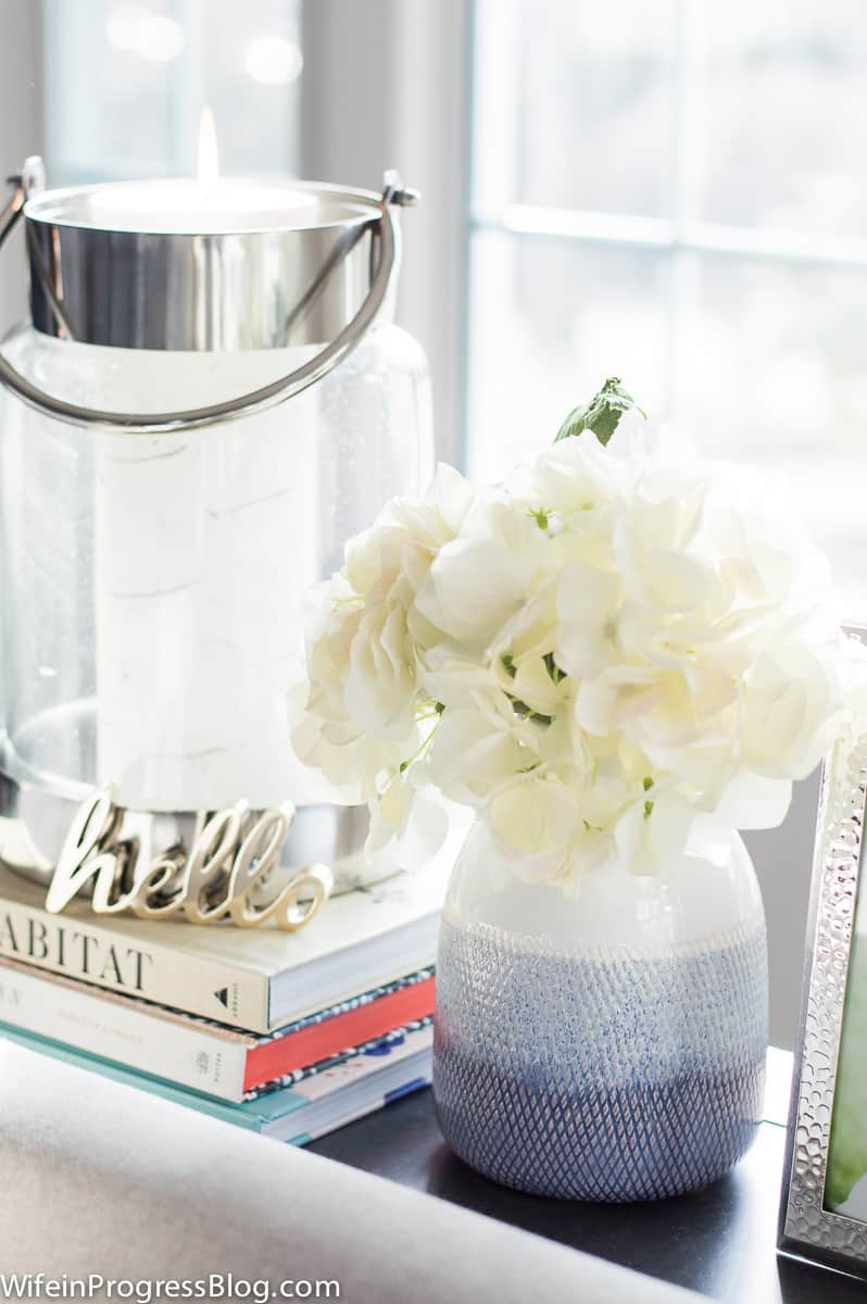 A light blue vase of white flowers, a clear glass lantern with a white candle, resting on a stack of books and a little decor trinket with the word \'hello\'