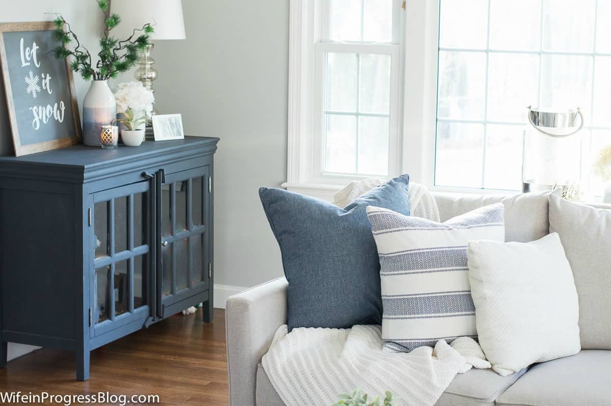 The end of a light grey sofa, with a group of blue and white pillows and a white throw, near a blue cabinet in the corner.