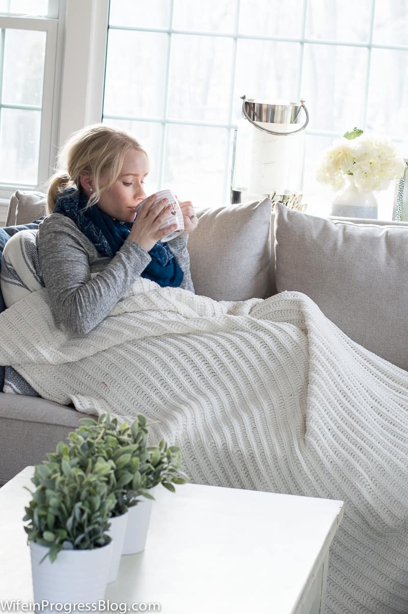 A woman, wearing a sweater and blue scarf, sitting on a sofa, covered with a white blanket, sipping from a mug 