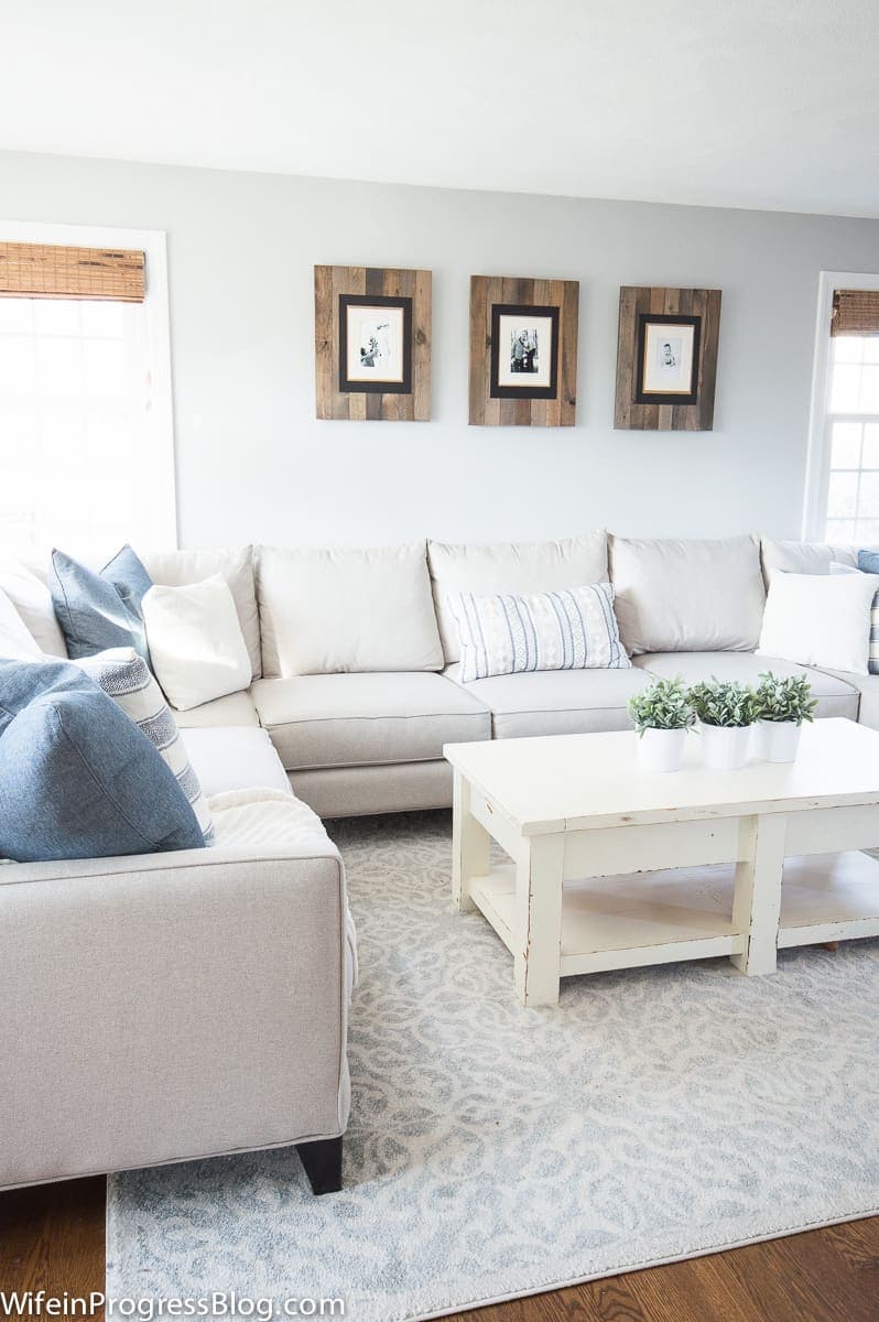 A living room with a light grey sofa, off-white wooden coffee table, 3 wooden photo frames on the wall and a large light blue area rug