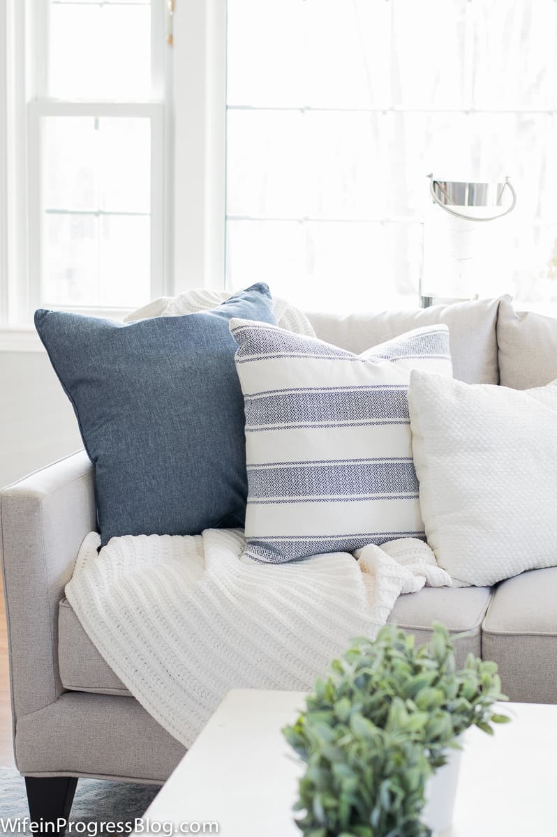 The end of the living room sofa, with a solid blue pillow, a horizontal striped blue and white pillow and a white pillow, with a white blanket tucked underneath