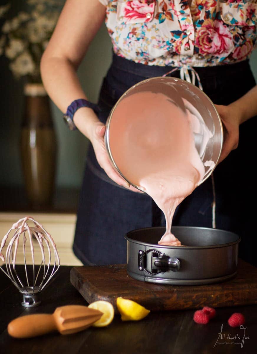 Pouring raspberry cheesecake mixture from the metal mixing bowl into a springform pan; meringue whisk nearby.
