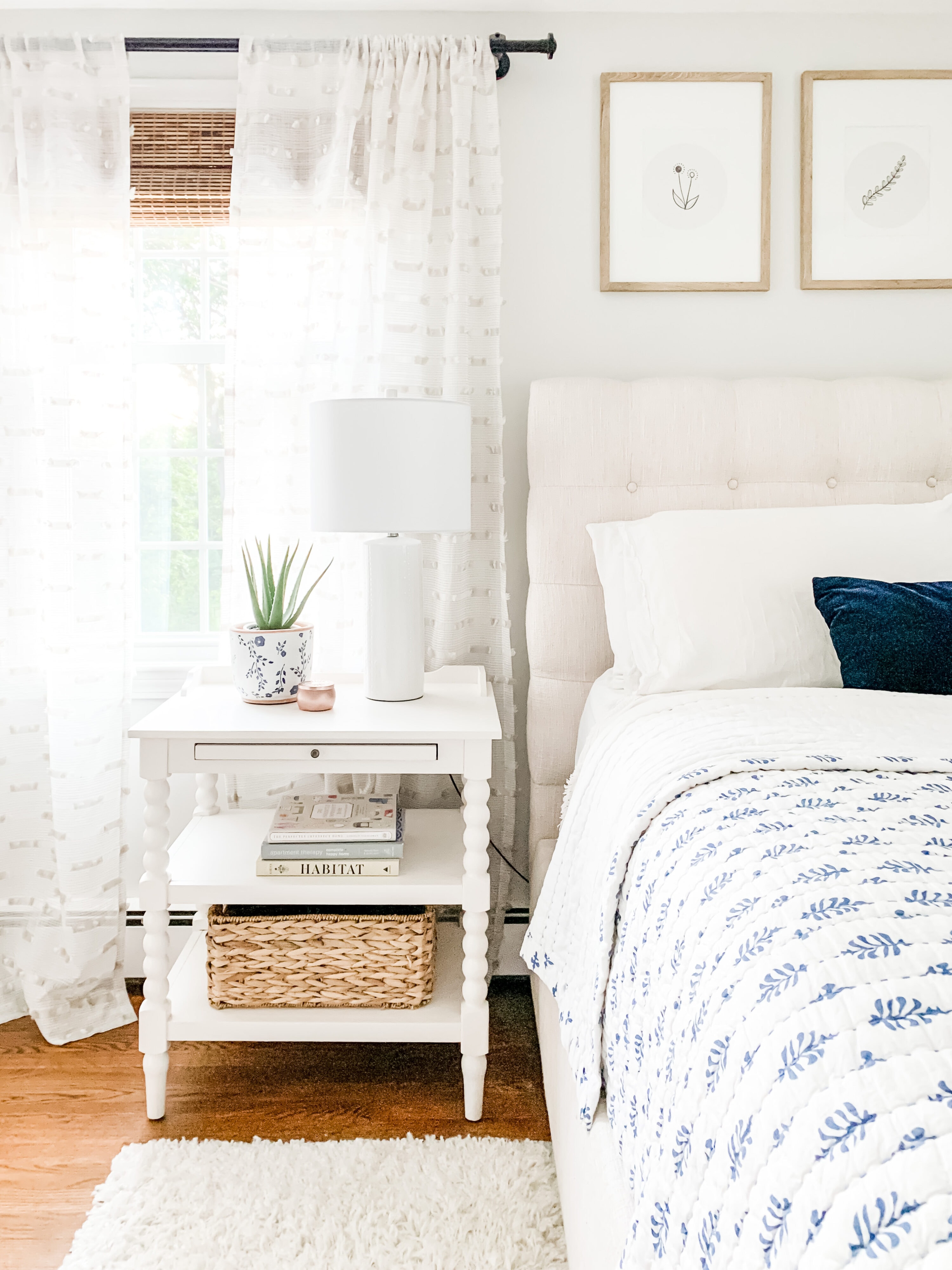 A bedroom with an off-white upholstered headboard, blue and white blanket, a white nightstand with various decor items and a window with white curtains