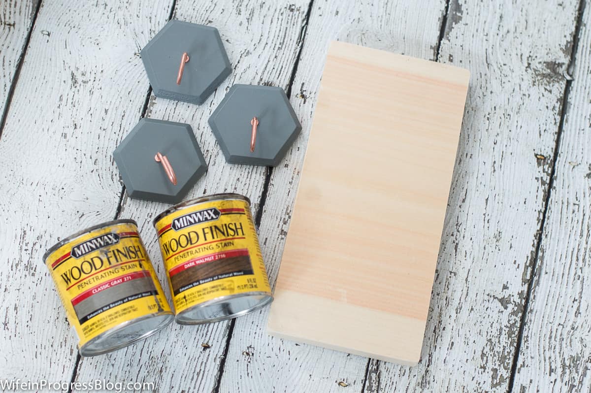 A natural plank of wood, 3 grey pieces of wood with hooks attached and 2 cans of stain nearby