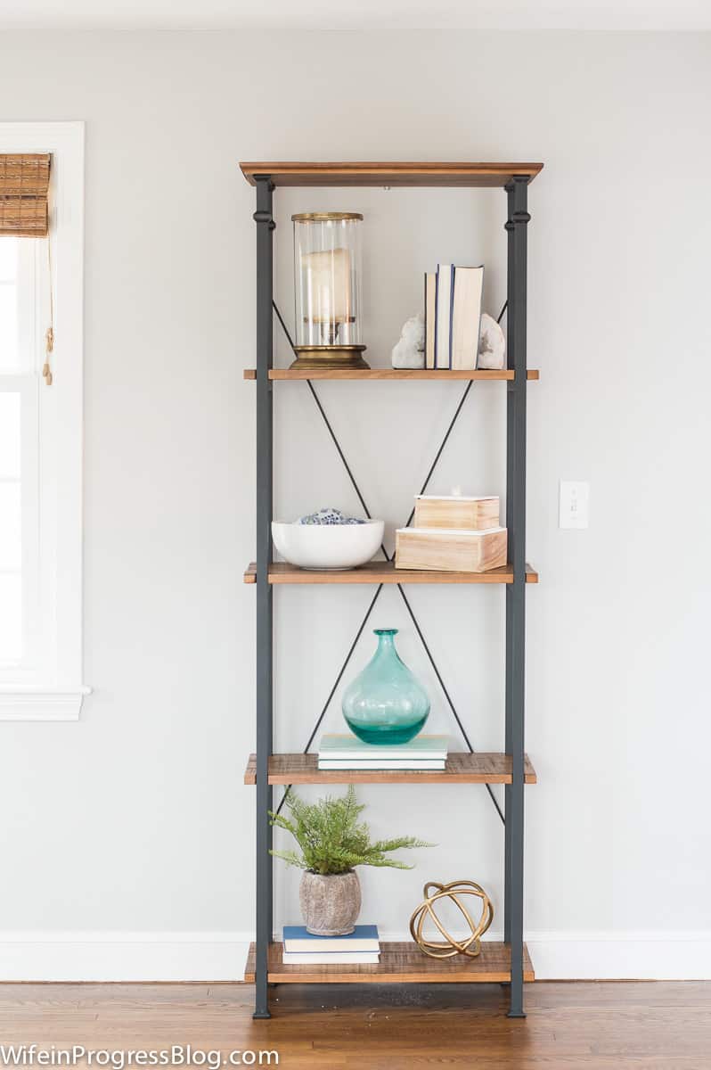 A four-tier bookcase with wooden shelving and metal frame, holding candles, books, decorative boxes and bowls and a few plants