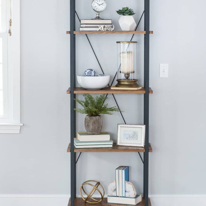A black metal bookcase with four wooden shelves, displaying stacks of books and various decor items in white, gold and blue tones