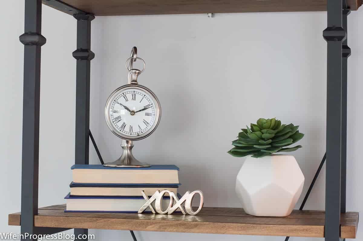 An antique silver clock, resting on a stack of books with the pages facing outward, near a potted plant on a wooden shelf