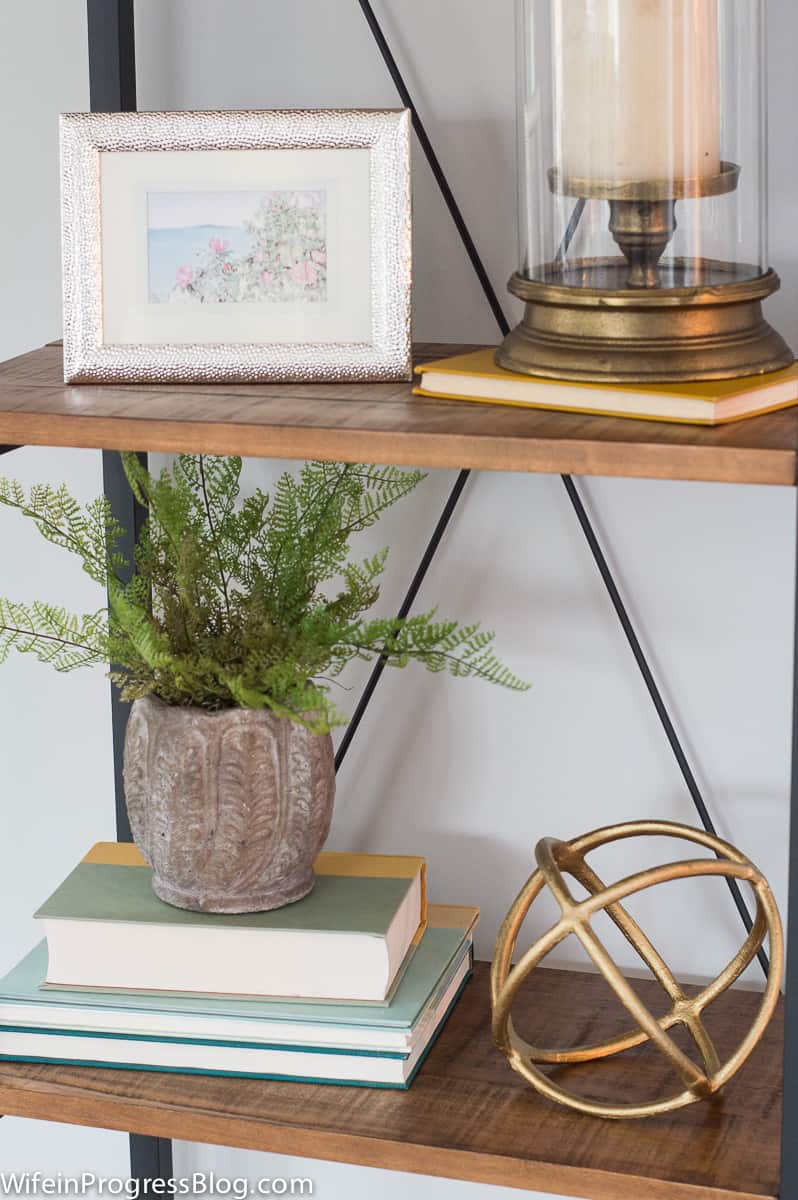 A bookcase with wooden shelf and metal frame, holding various decor items, including a plant, a lantern and a photo frame
