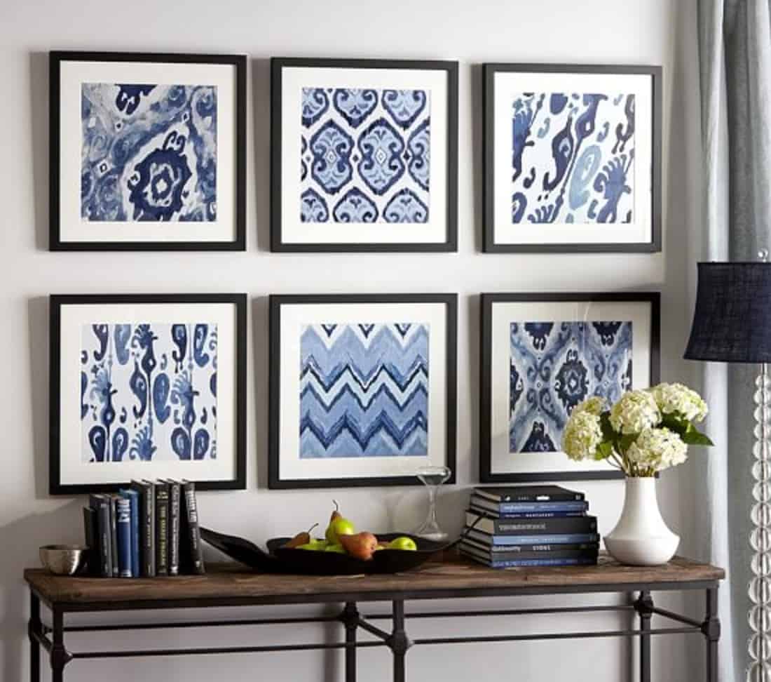 A group of six black frames displaying blue textile art above a console table containing a vase, books and tray of fruits