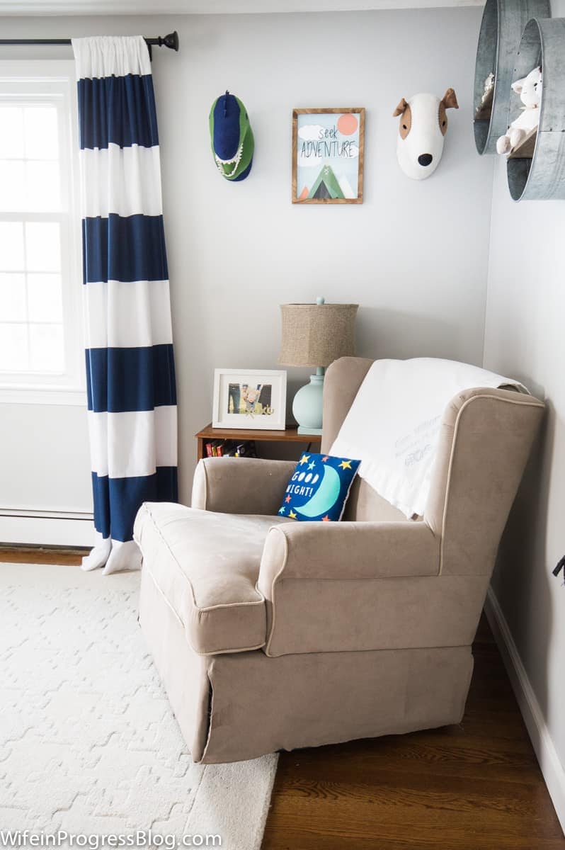 A nursery with beige, armchair rocker, blue and white curtains, and galvanized buckets mounted on the wall to hold stuffed animals