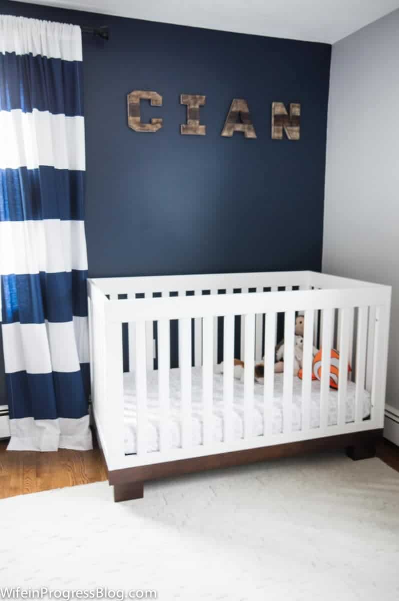 A white crib in front of a dark blue wall with the words "Cian" in wooden decor; a blue curtain with wide, horizontal blue and white stripes