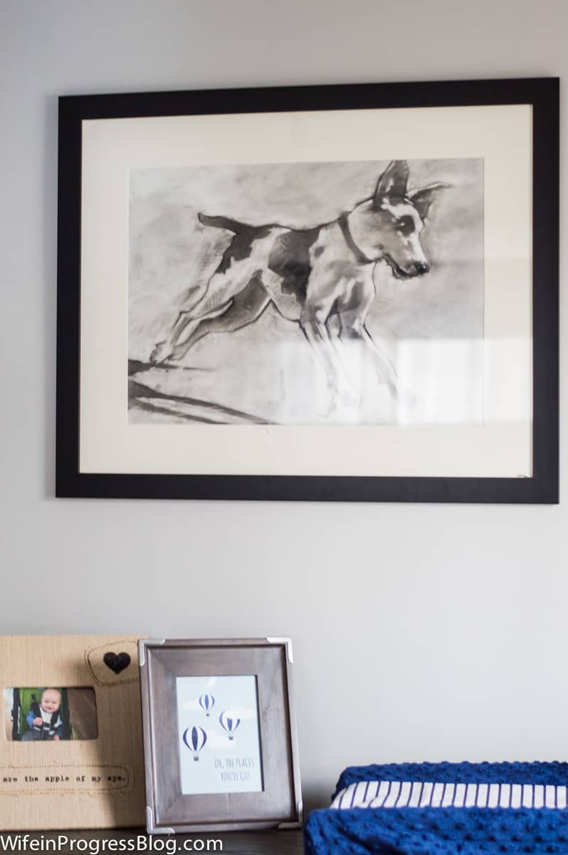 A large drawing of a dog on white matting and a dark frame, hung over a side table in a baby\'s room