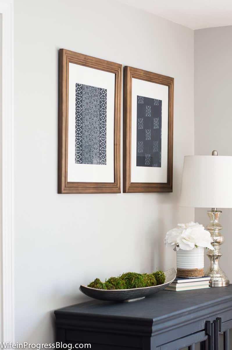 An angled view of the pair of framed textile art pieces above a blue table holding a silver  lamp and other decor items