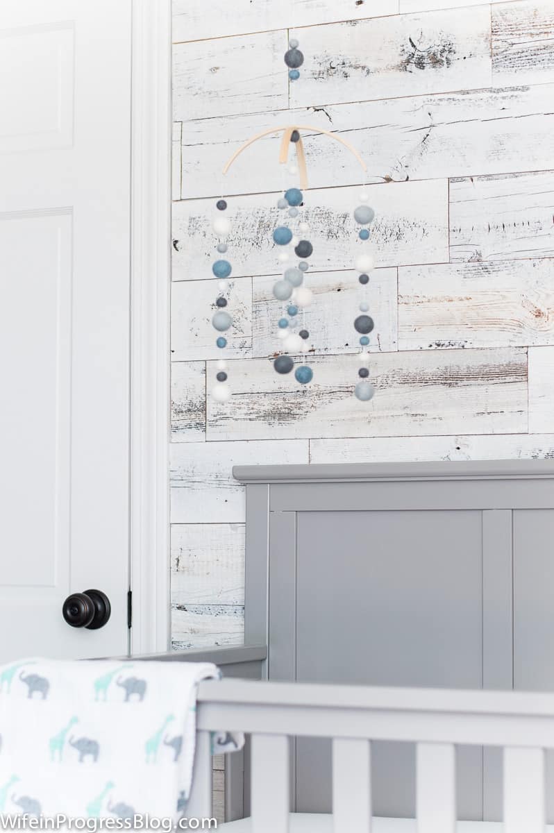 Baby boy nursery featuring blue and white mobile over grey crib set against reclaimed wood accent wall.