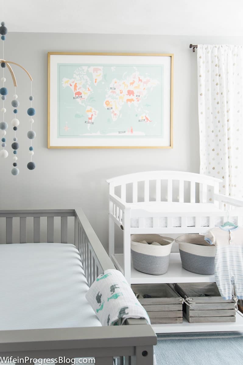 A nursery with a grey crib, blue and white mobile, white changing table and large world map on the wall in a wood frame