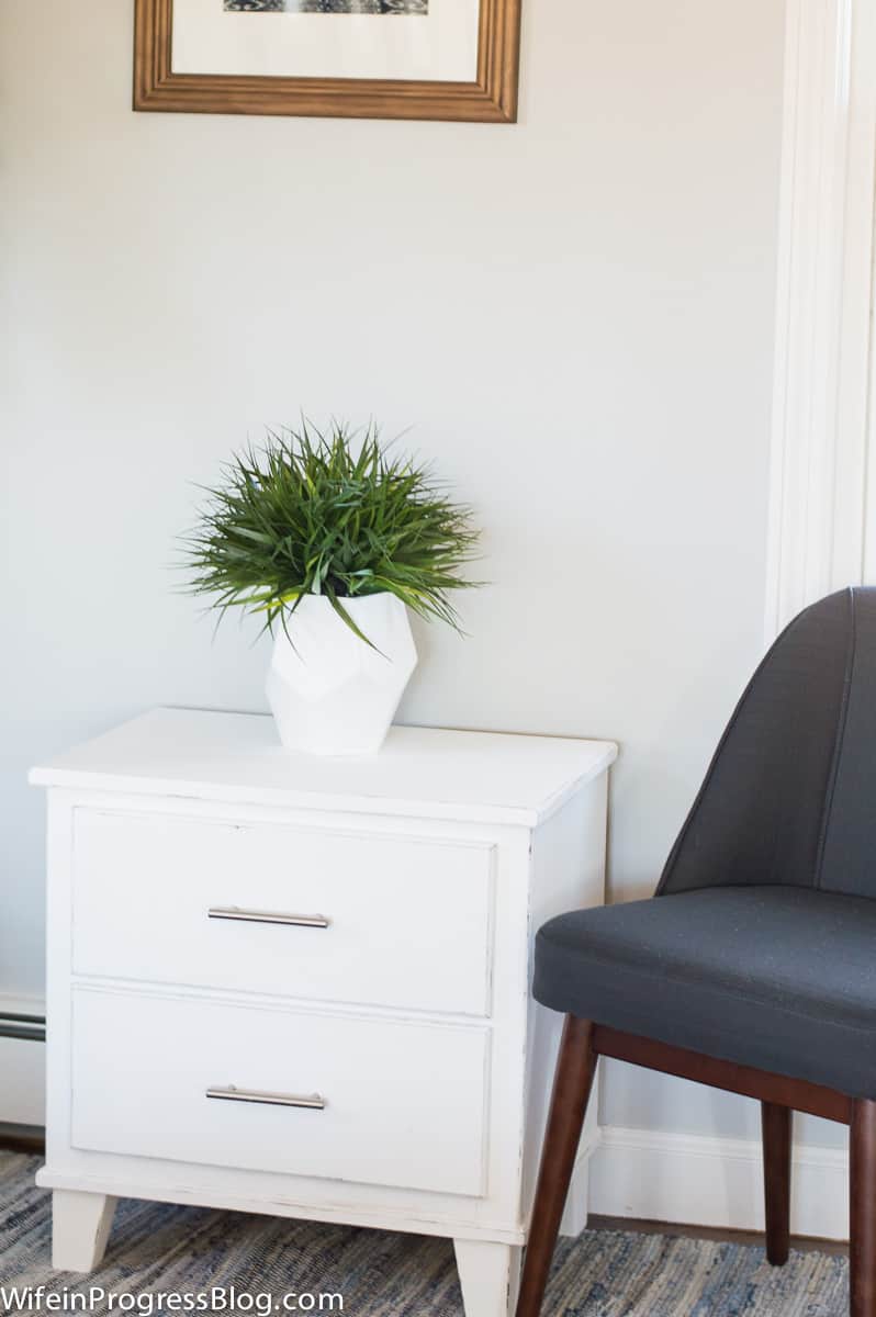 Nightstand, now white and distressed, holding an arrangement of greenery in a white vase, next to a blue accent chair