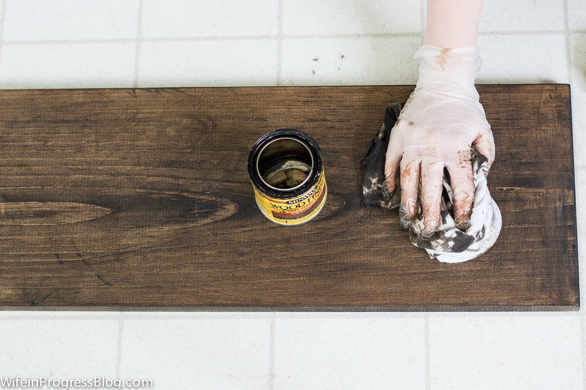 A person wearing gloves, apply dark wood stain to a plank of wood